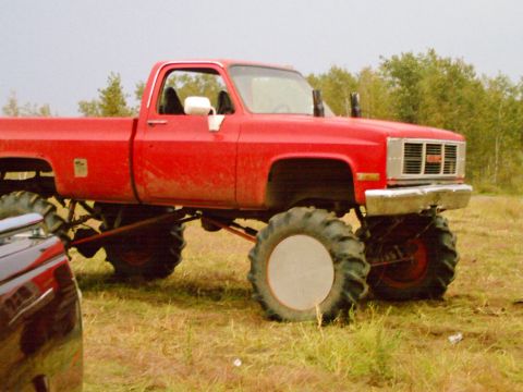 1976 Chevrolet Silverado 2500 - A heavily modified red pickup truck with massive tires sits in a grassy field, its GMC grille prominent.