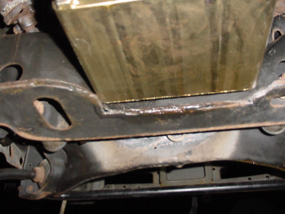Underside view of a gold-colored pan above a black crossmember with rusty suspension parts