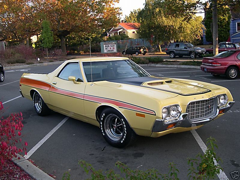 Yellow Ford Ranchero with orange stripe, parked in lot. Other cars and trees in background