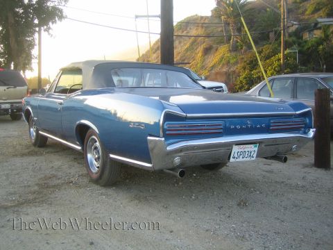 1968 Ford Mustang - A blue convertible is parked on a dirt lot with other vehicles nearby. The rear of the car shows its taillights and chrome bumper.