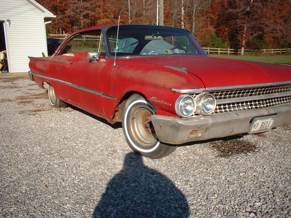 Faded red 1961 Ford Starliner hardtop parked on gravel, front-quarter view