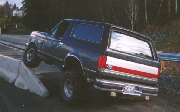 Blue full-size Ford Bronco with red stripe climbing over a concrete barrier