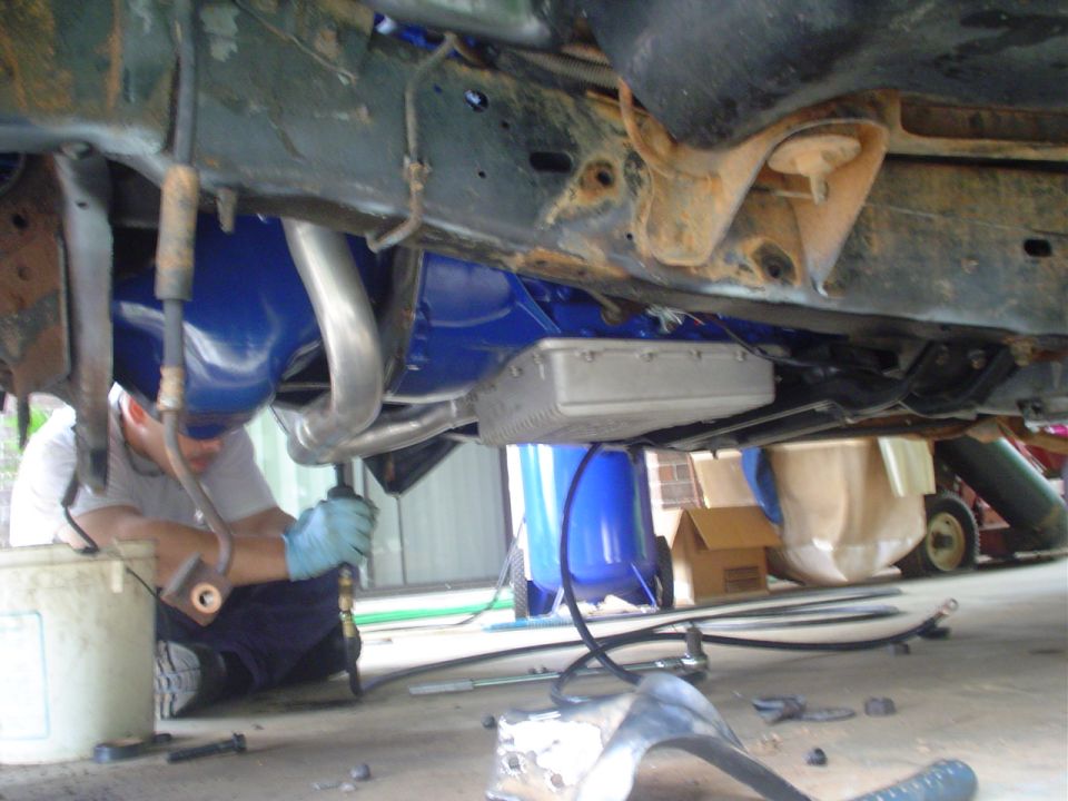 Underside of raised truck showing blue V8 engine, headers and trans pan while a person works below