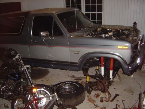 1986 Ford Bronco - 1986 Ford Bronco is on jack stands with front suspension removed for work. A motorcycle is parked nearby.