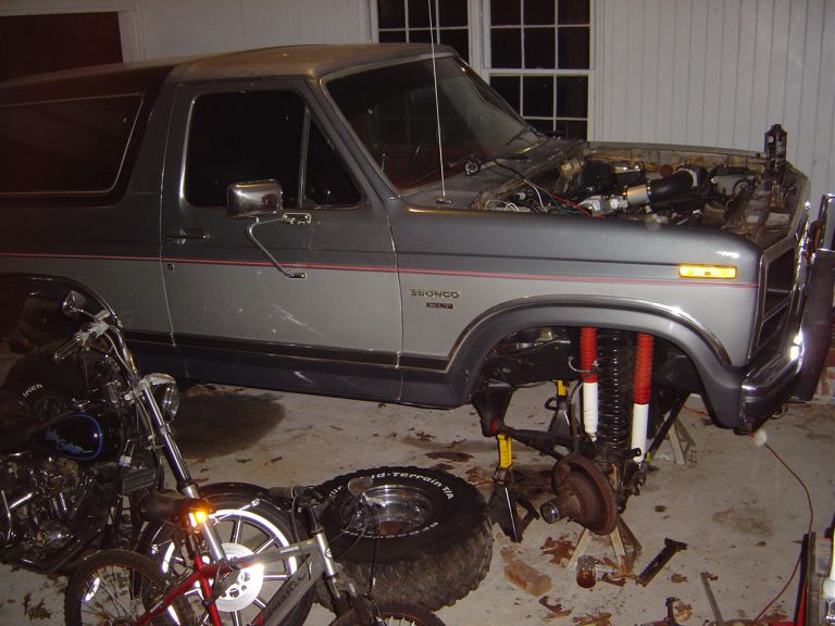 1986 Ford Bronco with hood open and front end disassembled, parked next to a motorcycle