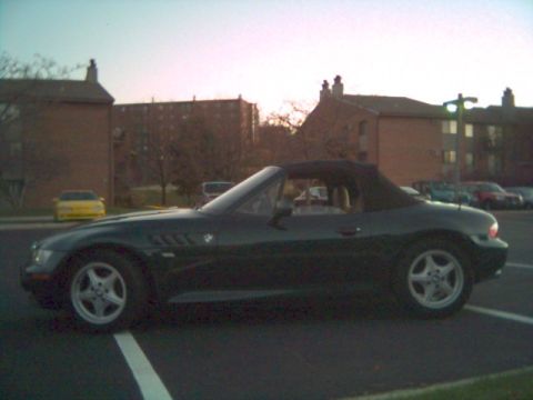 1996 BMW Z3 - A dark convertible sports car is parked in a lot, showing its side profile against a backdrop of buildings.