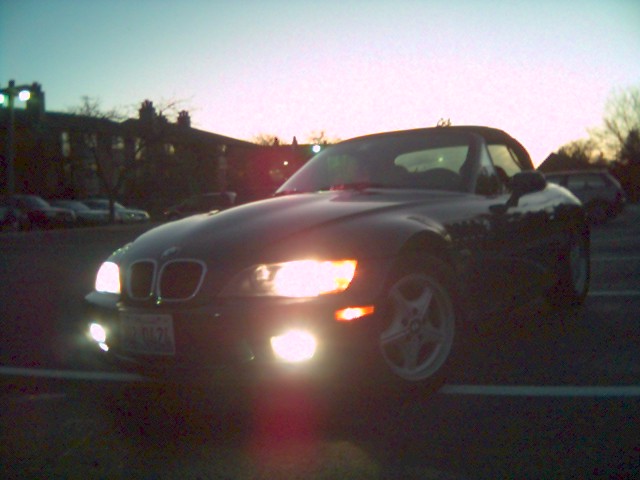 Dark BMW Z3 roadster at dusk with headlights and fog lights on in a parking lot