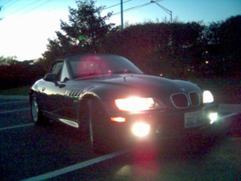 1996 BMW Z3 - A dark BMW Z3 convertible is parked in a lot as dusk settles, its headlights illuminating the scene.