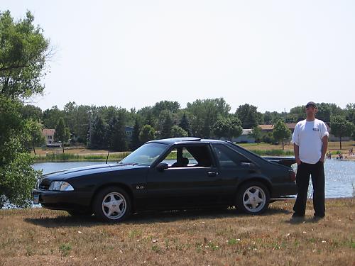 Black Fox-body Mustang LX 5.0 hatchback parked on grass beside a lake, man standing at rear