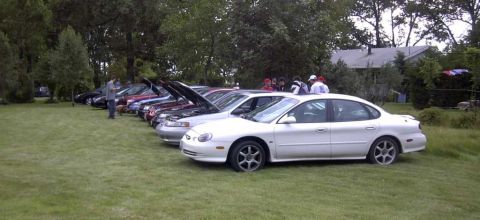 1997 Ford Taurus - A line of cars, including a white 1997 Ford Taurus, are parked on a grassy lawn during an outdoor gathering.