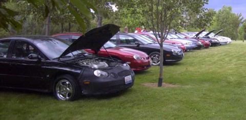 1997 Ford Taurus - A line of cars, including a black 1997 Ford Taurus with its hood open, are parked on a grassy lawn.