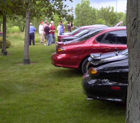 1997 Ford Taurus - Two sedans, a red and a black one, are parked on a grassy area with trees and people in the background.