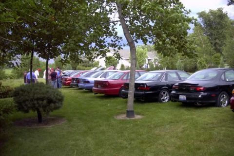 1997 Ford Taurus - A line of sedans, including a black 1997 Ford Taurus, are parked on a grassy lawn under trees.
