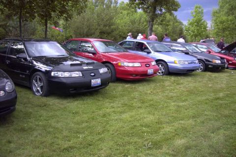 1997 Ford Taurus - Several sedans, including a black 1997 Ford Taurus, are parked on a grassy lawn with trees in the background.