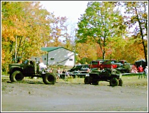 1976 Chevrolet Silverado 2500 - Off-road vehicles are gathered in a wooded area with a building and tents visible in the background.