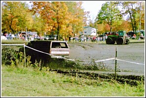 1976 Chevrolet Silverado 2500 - A van navigates a muddy track during an event, with a tractor visible in the distance.