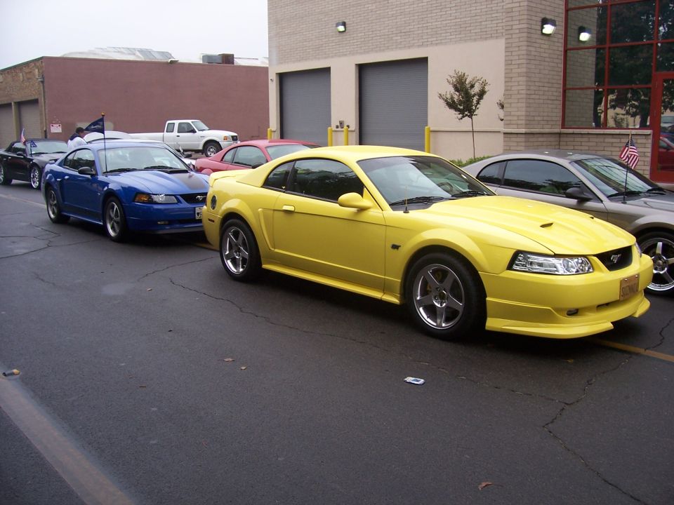 Yellow 2002 Ford Mustang in front of a blue one and a gray one, parked on asphalt