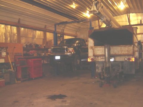 1992 Ford Bronco - A black Ford F-Series truck is raised on a lift inside a garage, with a dump truck parked nearby.