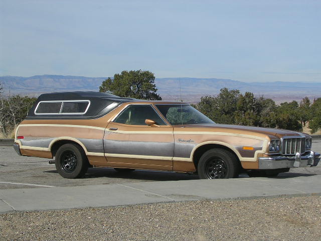1975 Ford Ranchero with a black camper shell parked on a paved surface with mountains in the background
