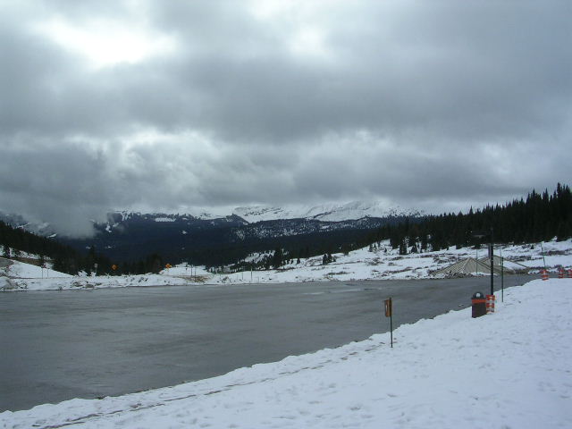 snowy landscape with a frozen lake, pine trees, and mountains under a cloudy sky. A trash can is visible on the right