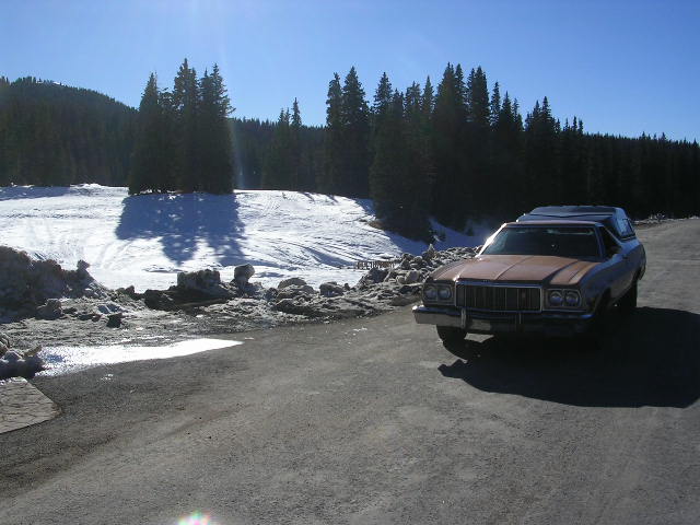1975 Ford Ranchero with a camper shell parked on asphalt with a snowy hillside and trees in the background
