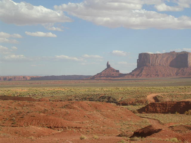 Desert landscape with Monument Valley rock formations and a dirt road, under a cloudy sky. Supermotors.net logo visible