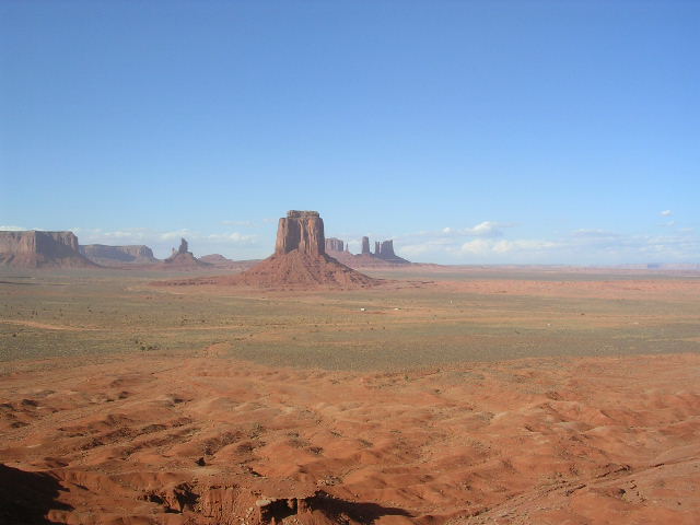 Monument Valley with iconic buttes under a clear blue sky. Foreground shows red desert terrain