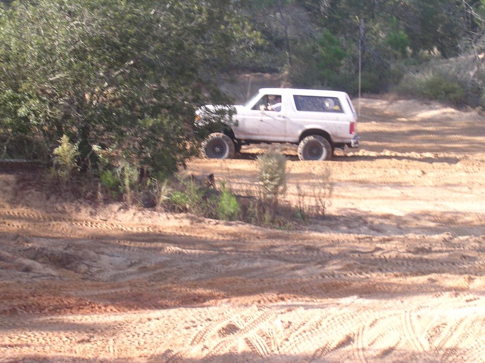 White Ford Bronco driving on a sandy trail with trees to the left