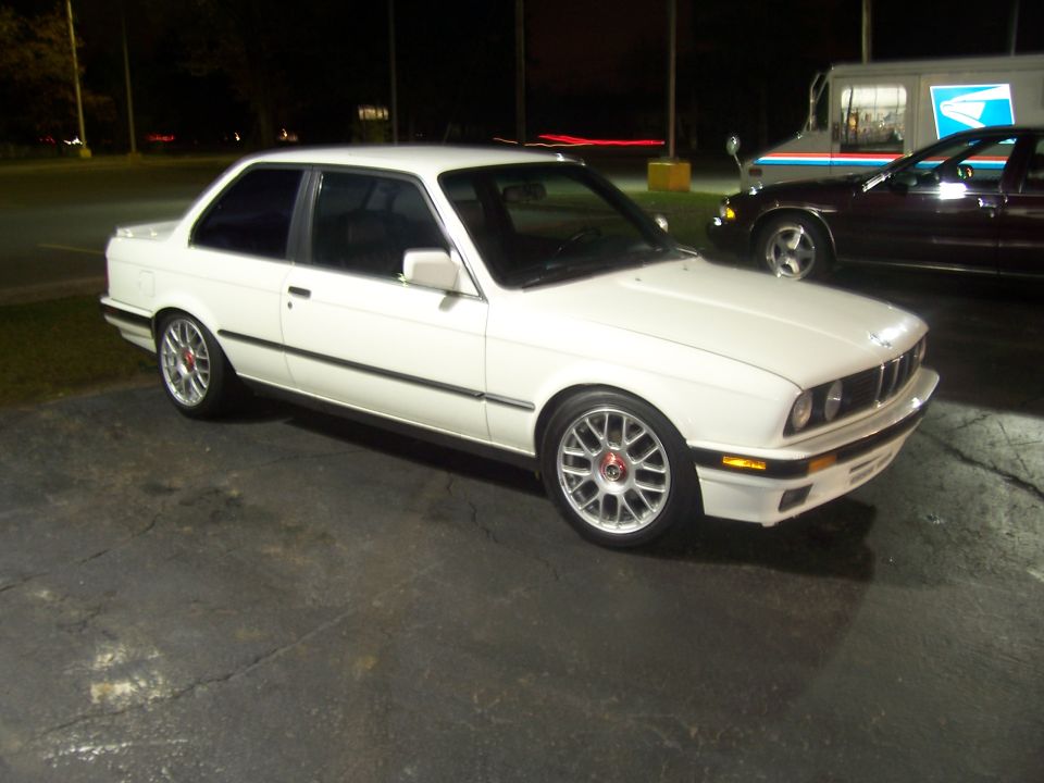 White BMW E30 coupe with aftermarket wheels parked on asphalt, with a USPS truck and another sedan behind it