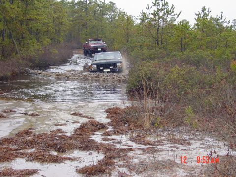 2004 Jeep Wrangler - Two off-road vehicles navigate a muddy, water-filled trail surrounded by trees.