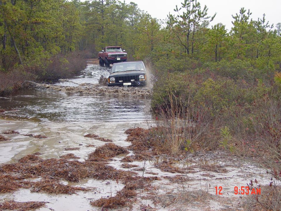 Jeep Cherokee XJ driving through water, red pickup truck behind it