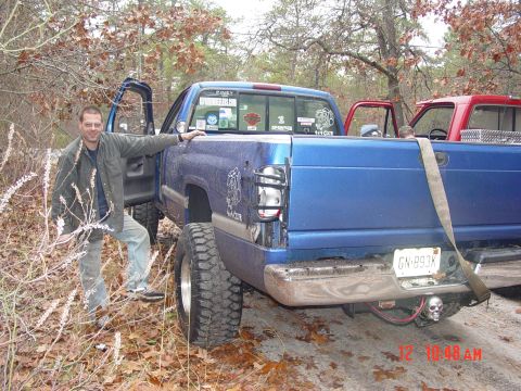 2004 Jeep Wrangler - A man stands beside a lifted blue pickup truck, with another red truck visible behind it.