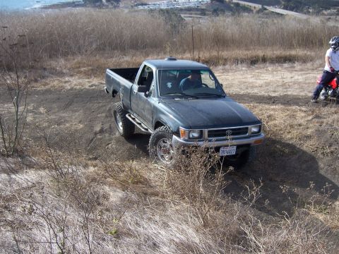 1995 Toyota Hilux - 1995 Toyota Hilux pickup truck navigating an off-road trail with a dirt bike rider nearby.