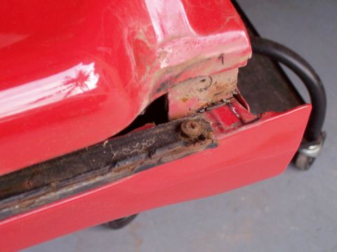1978 Ford Bronco - Tight close-up of a red truck's lower rear body corner with the bedside or quarter panel pulled away, exposing a rusty, dirt-caked mounting…