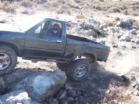 1995 Toyota Hilux - A pickup truck is seen off-roading on a rocky hill, kicking up dust as it maneuvers.