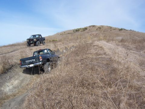 1995 Toyota Hilux - Two Toyota pickup trucks navigate a steep, dry, grassy incline under a clear blue sky.