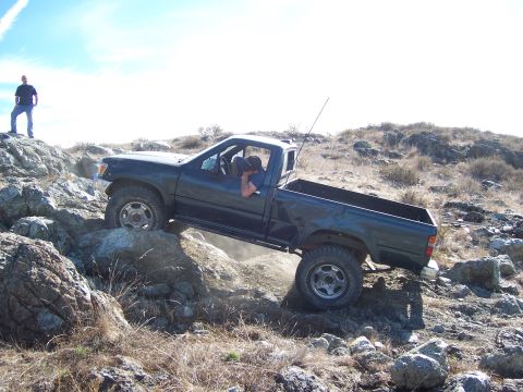 1995 Toyota Hilux - A dark green pickup truck is shown off-roading over rocks, with a person standing on a boulder in the background.