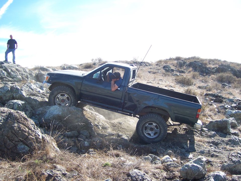 Dark green pickup truck navigating a rocky incline. The truck has a single cab and appears to be from the early 1990s