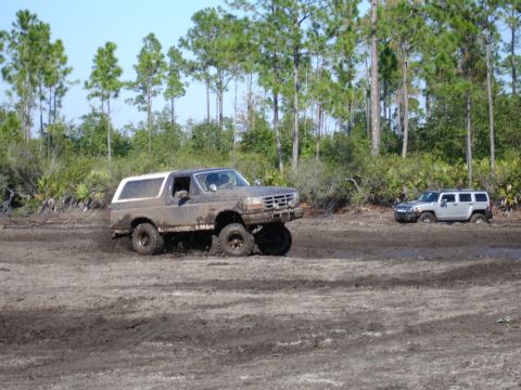 1989 Ford Bronco - A muddy 1989 Ford Bronco navigates a challenging off-road terrain, with another vehicle partially submerged in the mud behind it.