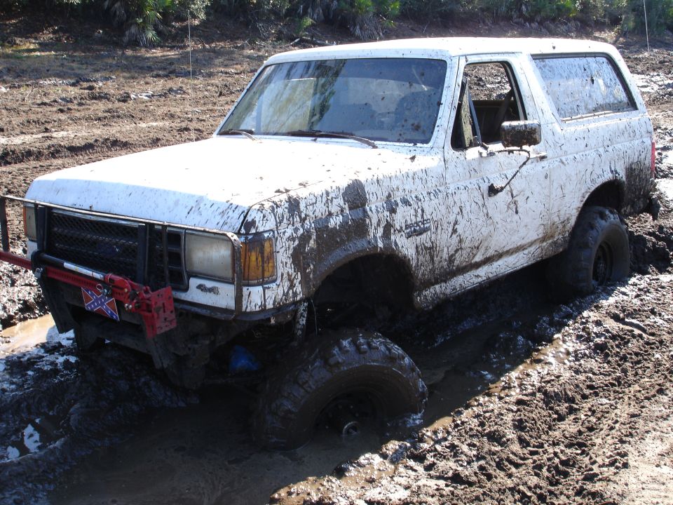 Mud-covered white Ford Bronco with a red jack attached to the front bumper, stuck in a muddy pit