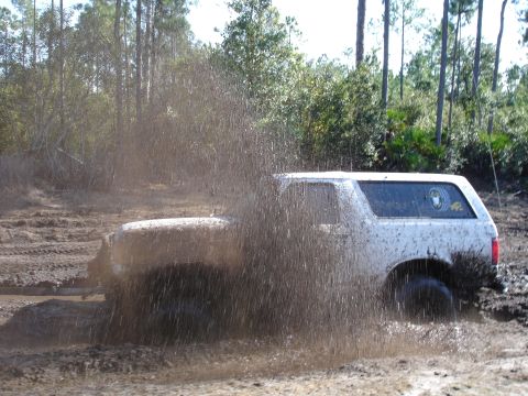 1989 Ford Bronco - A white SUV navigates a muddy track, kicking up a large spray of dirt and water.