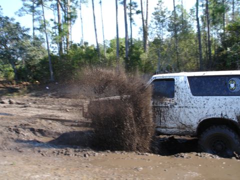1989 Ford Bronco - A white SUV navigates a muddy trail, creating a large splash as it drives through the mire.