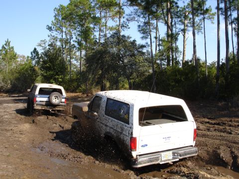1989 Ford Bronco - Two Ford Broncos navigate a muddy off-road course with trees in the background.