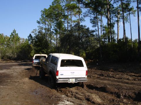 1989 Ford Bronco - Two white SUVs navigate a muddy track through a wooded area on a sunny day.