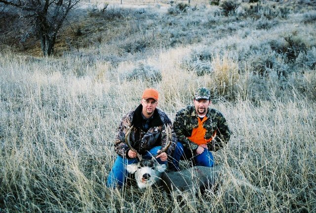 Two men in camouflage kneel in dry grass with a deer carcass between them. One man holds the deer's antlers