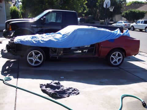 1971 Ford Mustang - A red car body, partially covered by a blue tarp, sits in a driveway next to a black pickup truck.