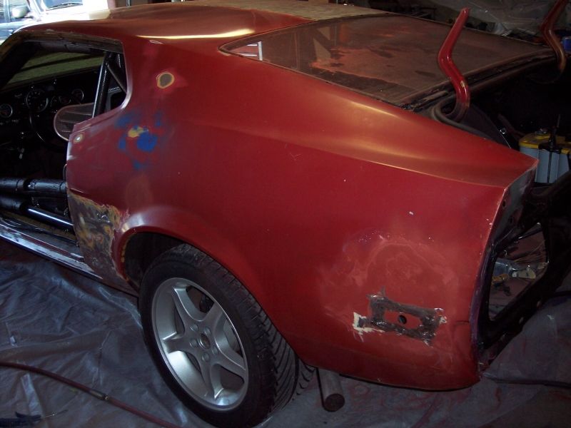 car body with red paint, primer, and rust spots. Trunk open, showing red supports. Rear quarter panel has rust and weld repairs