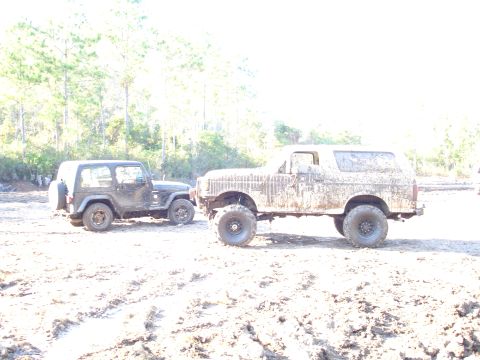 1989 Ford Bronco - Two off-road vehicles, a Ford Bronco and a Jeep Wrangler, are covered in mud after driving through a sandy, wooded area.