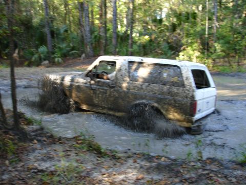 1989 Ford Bronco - A white 1989 Ford Bronco navigates a muddy trail, kicking up water and mud.
