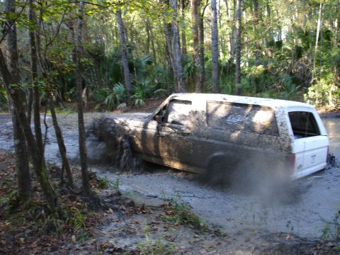 1989 Ford Bronco - A white 1989 Ford Bronco navigates a muddy trail, kicking up water and dirt.